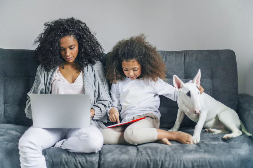 A mother, her daughter and their dog lounge on a couch. The woman is using a laptop, while the girl browses her tablet and pets the dog.