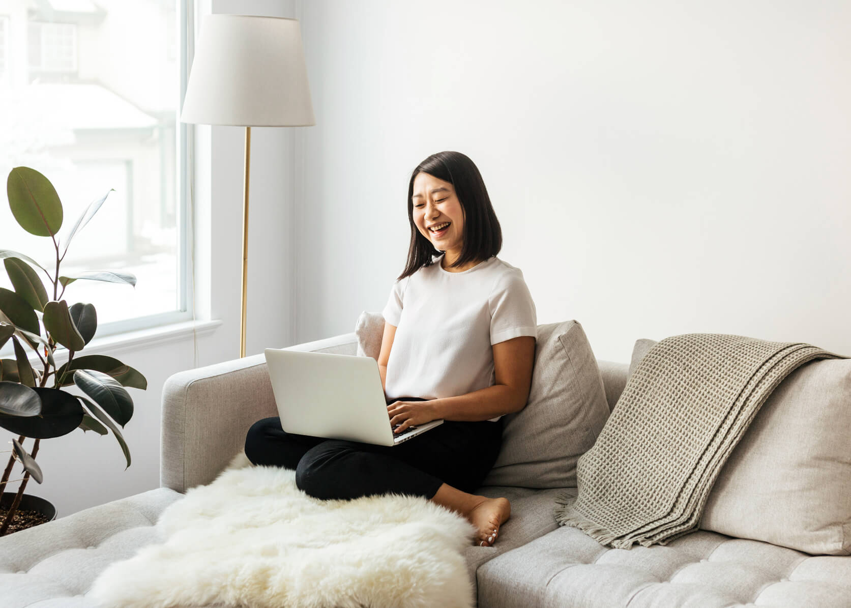 A smiling woman sits on her couch with an open laptop