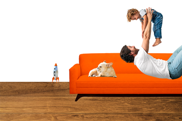 A man lying on a couch next to the family dog holds a happy toddler above his head